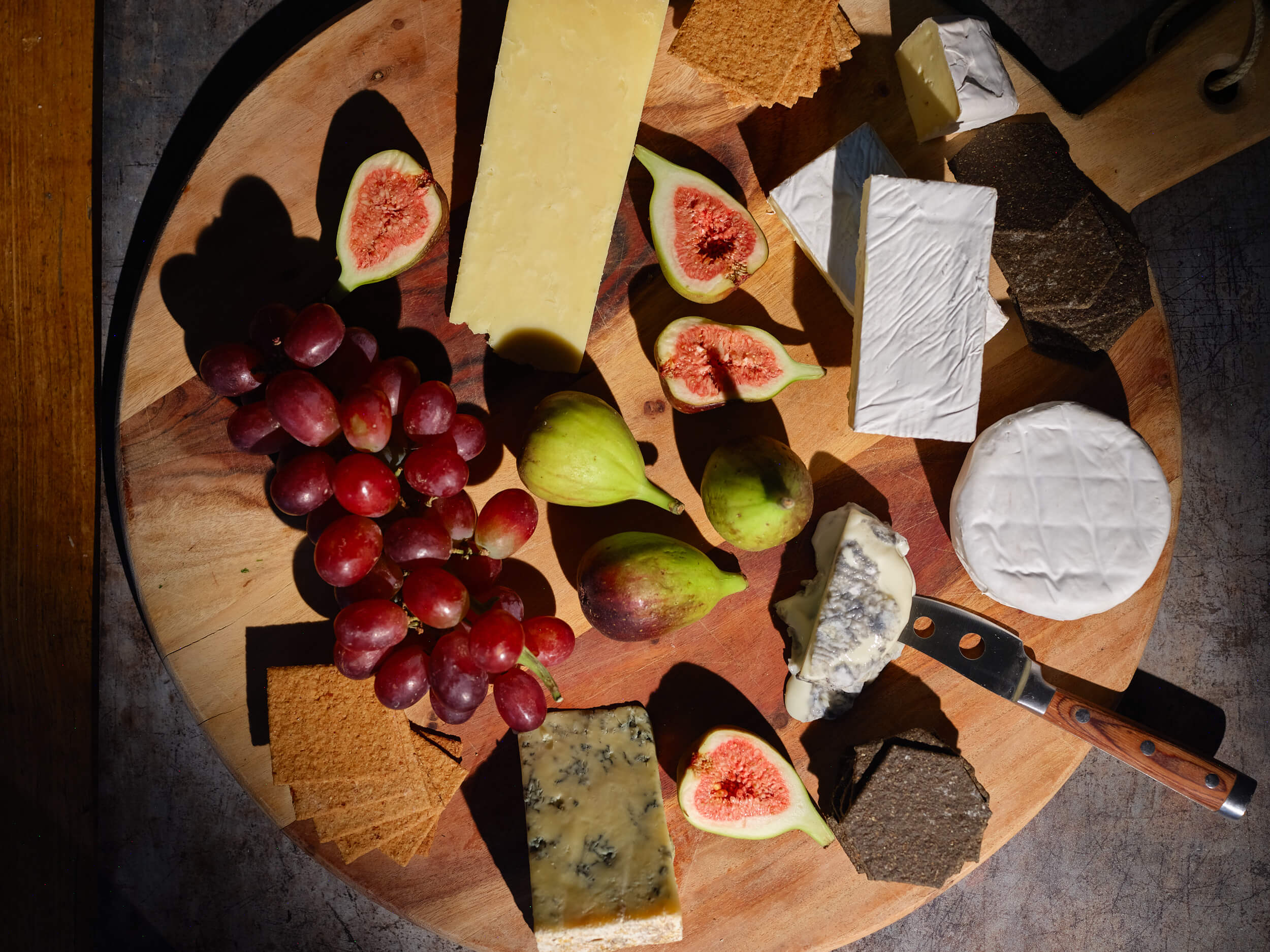 Rounded rustic cheeseboard with a variety of cheese and fruit, alongside a cheese knife.