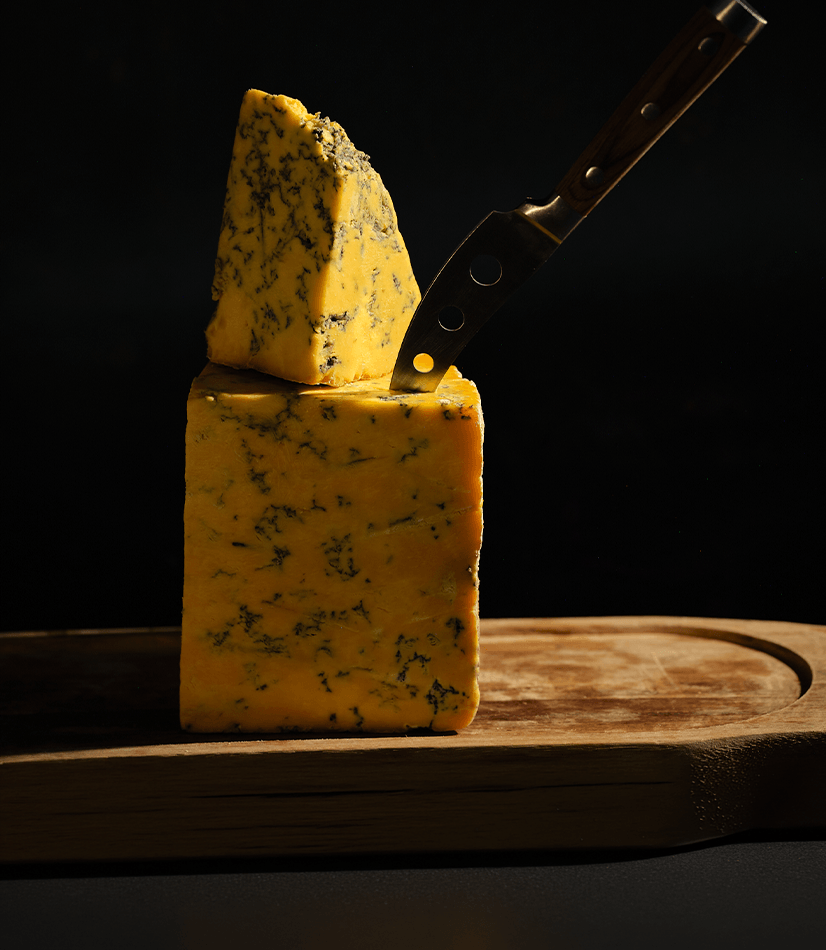 Yarlington Blue Cheese from Longmans, presented on a rustic board creatively stacked. A knife is placed in the main block.